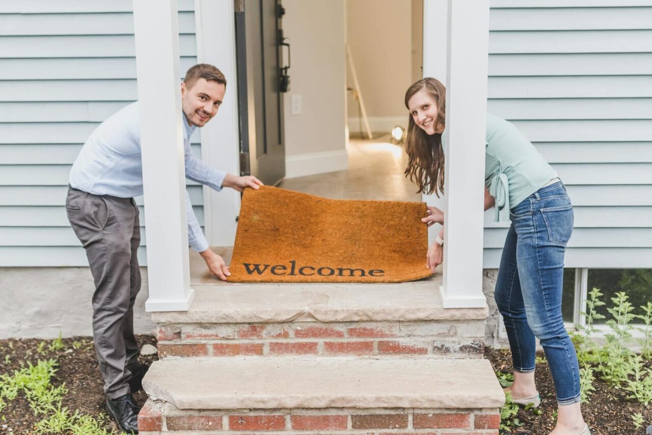 Couple smiling while holding a 'welcome' mat at their home's front door, symbolizing a warm greeting.