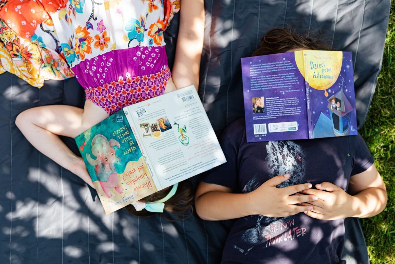 Two children lie outdoors on grass, faces covered by books, enjoying a sunny day.
