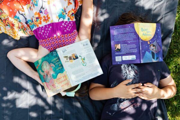 Two children lie outdoors on grass, faces covered by books, enjoying a sunny day.